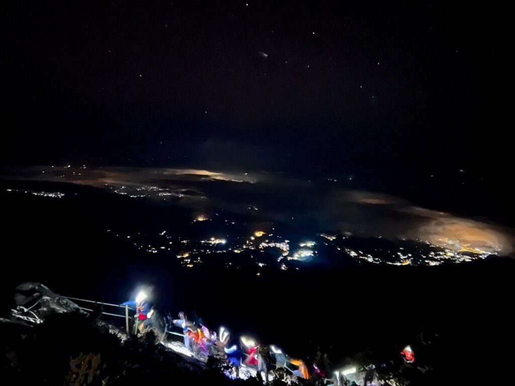 Hikers climbing Mount Kinabalu summit push at predawn with headlamps, forest trail, distant city lights, and starry sky. © SlowingwithT.com