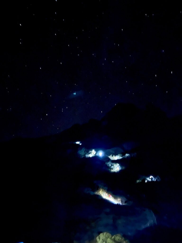 Hikers’ headlamps forming a glowing light trail on a dark predawn ascent of Mount Kinabalu with mountain silhouette. © SlowingwithT.com
